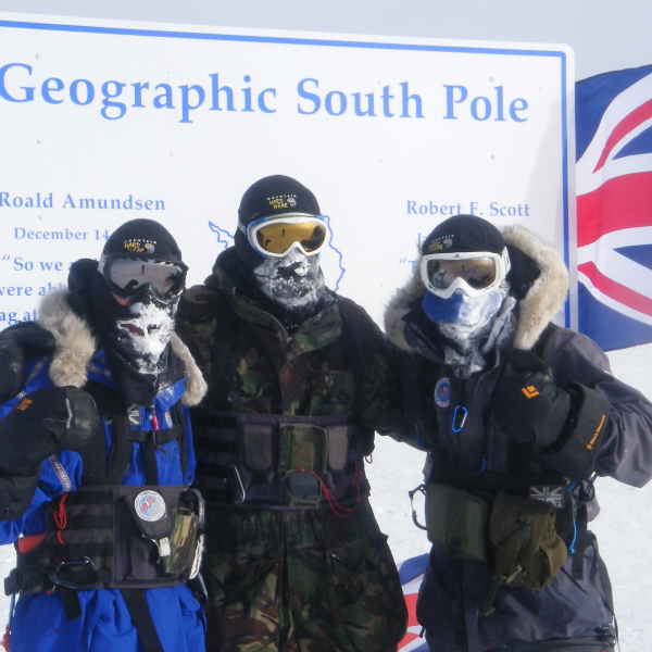 Three people wearing full cold weather gear stand in front of a sign reading 'Geographic South Pole'.