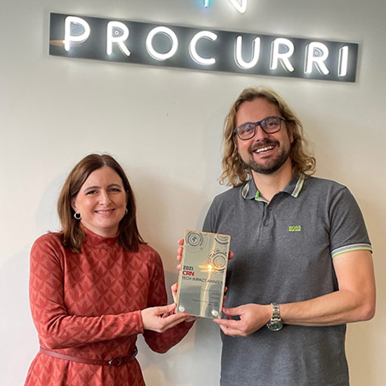 Two Procurri employees hold up an award plaque below a sign that reads 'Procurri'.