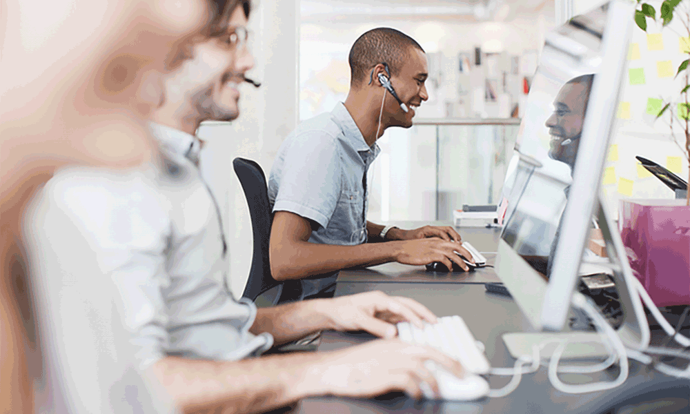 mws-3-2 A row of three people sit in front of computers on a desk. The third person is in focus and is smiling at the screen.