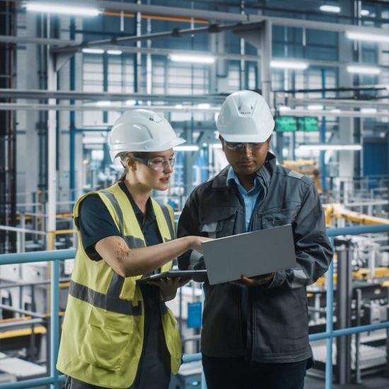 A male and female both dressed in white hard hats against an industrial backdrop look at a laptop and tablet device.
