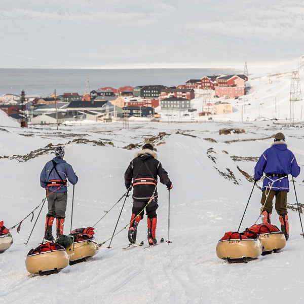 Three people skiing along snow dragging bags behind them head off camera to a small village of colourful buildings in the distance.