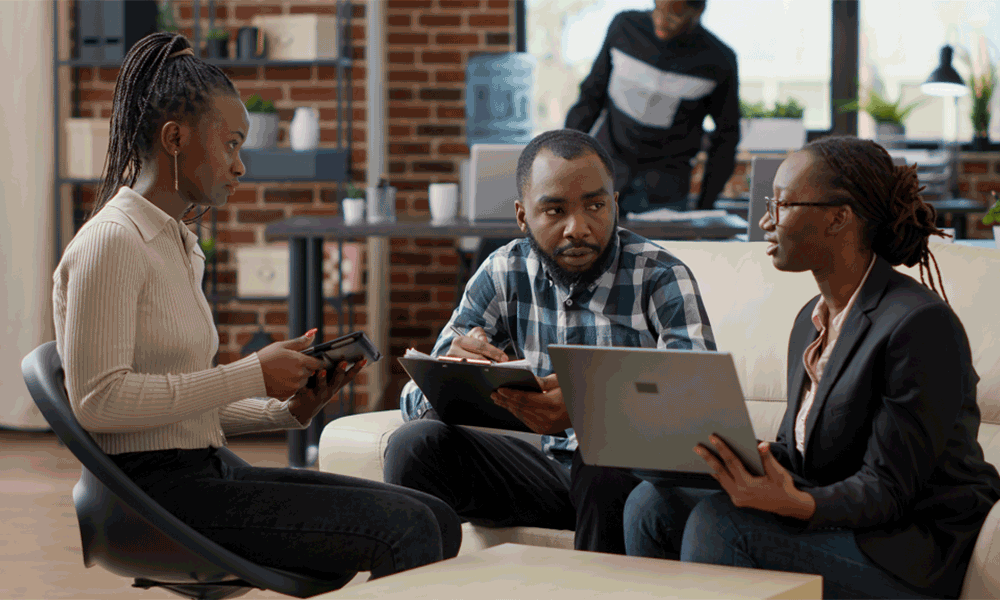 ps-3 Three adults are sitting on a sofa and stool chatting against a modern office backdrop. Each holds a device; one smartphone, one tablet, one laptop.